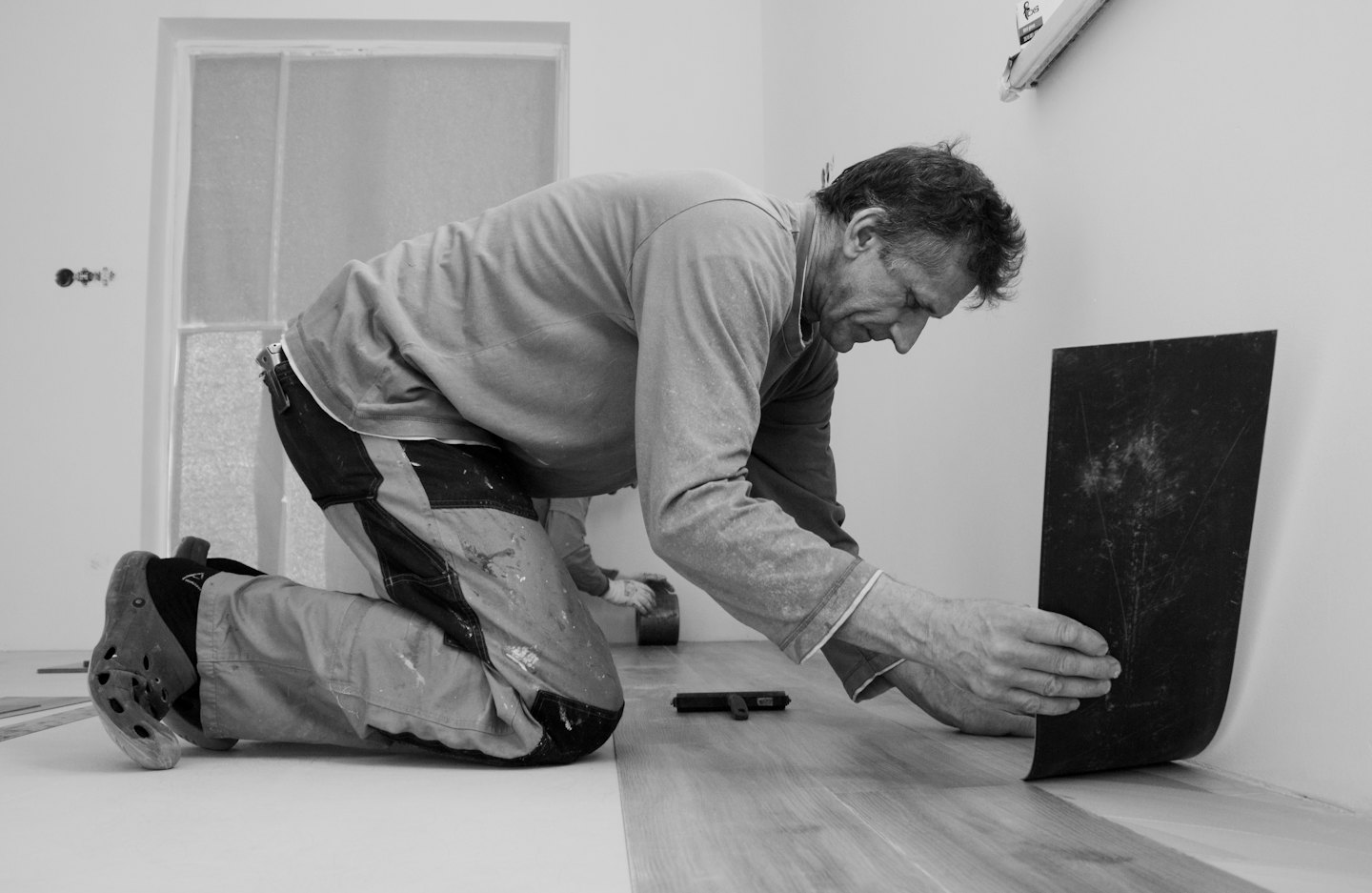 Man installs new wooden-look flooring in a white-walled room.